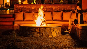 An outdoor seating area with a fire pit, surrounded by palm trees and a building with arched windows.