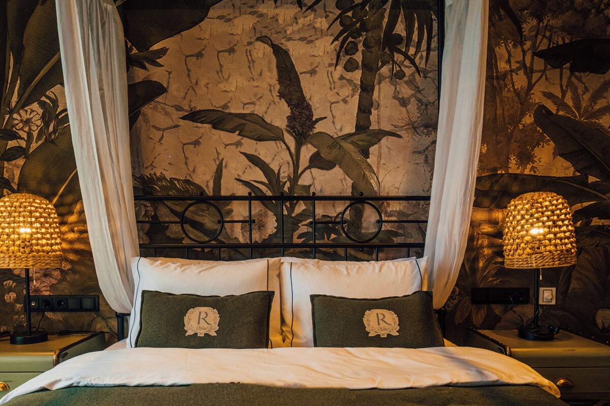 Una cama con dosel, dos almohadas con fundas con monogramas y un mural de follaje en la pared.