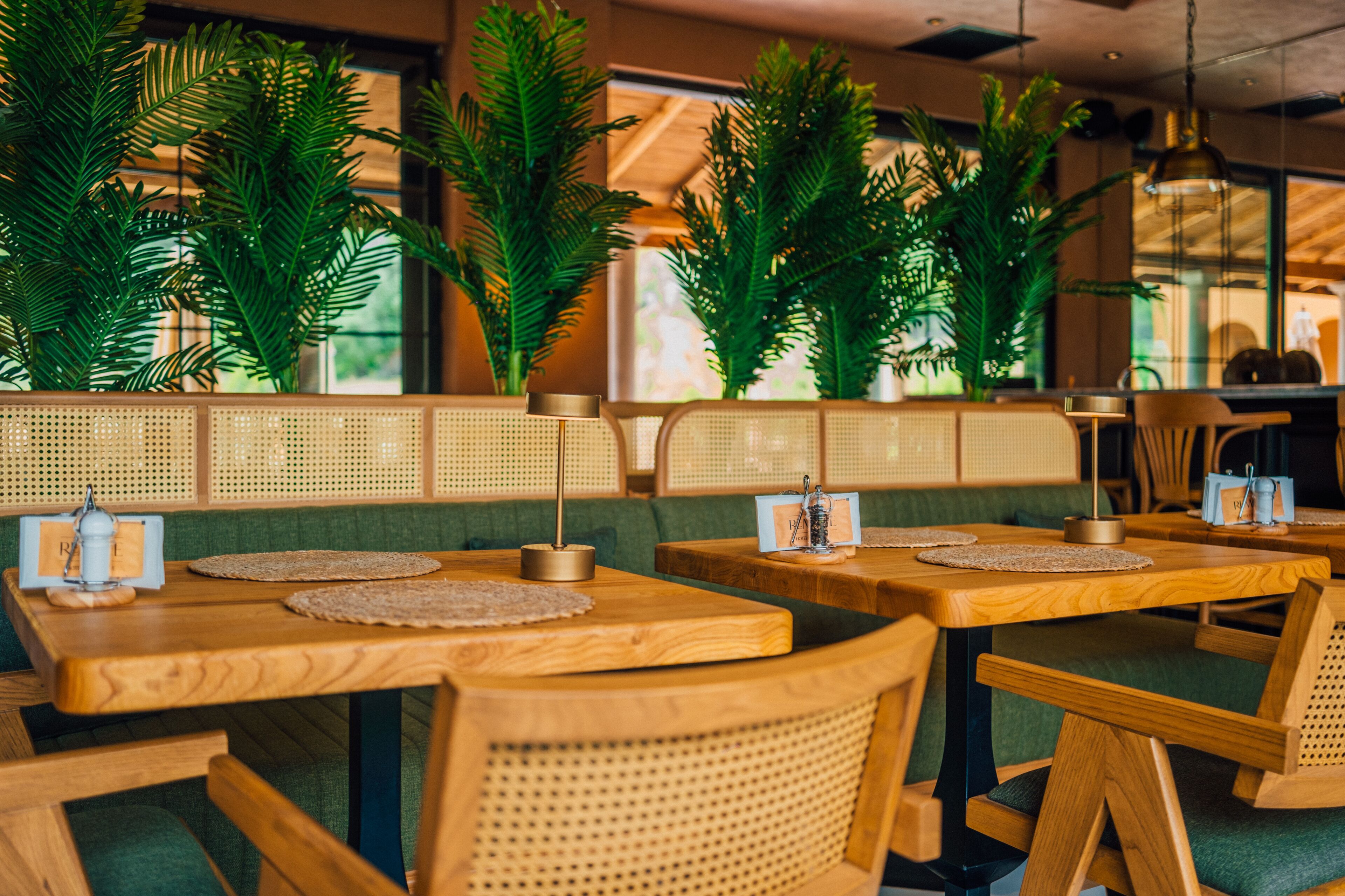 Ein Restaurant mit Holztischen und grün gepolsterten Sitzbänken, umgeben von hohen Grünpflanzen.