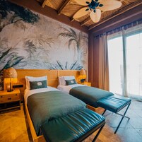 Habitación de hotel con dos camas, un mural grande de palmeras, un ventilador de techo y vistas al exterior a través de una ventana.
