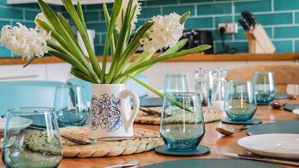 Ein Esstisch ist gedeckt mit blauem Glas, einer weißen Blumenkanne und einer gewebten Tischmatte.