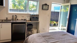 Eine kleine Küche mit Kühlschrank, Toaster, Wasserkocher und Backofen. Ein Fenster mit Jalousien, eine Uhr und eine Tür nach draußen.