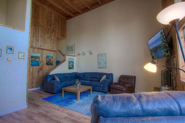 Een woonkamer met een houten plafond, een blauwe bank, een salontafel en een aan de muur bevestigde televisie.