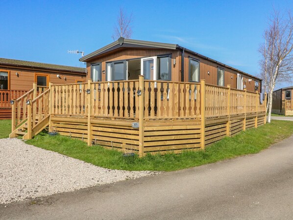 A wooden mobile home with a fenced deck, a staircase, and a gravel driveway. - Birch Lodge (Carnforth)