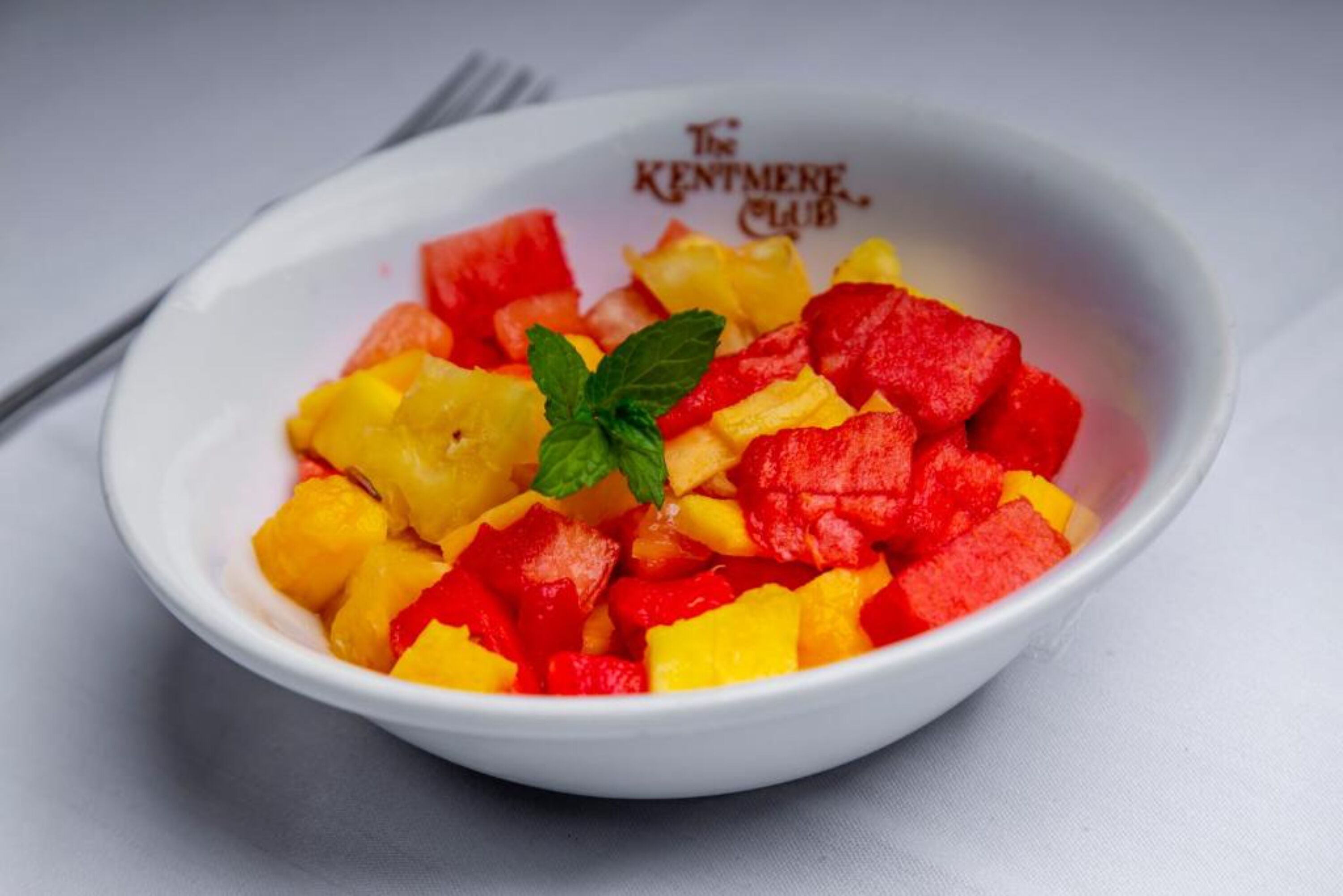 A white bowl with diced red and yellow fruit, garnished with a mint leaf, on a white surface.