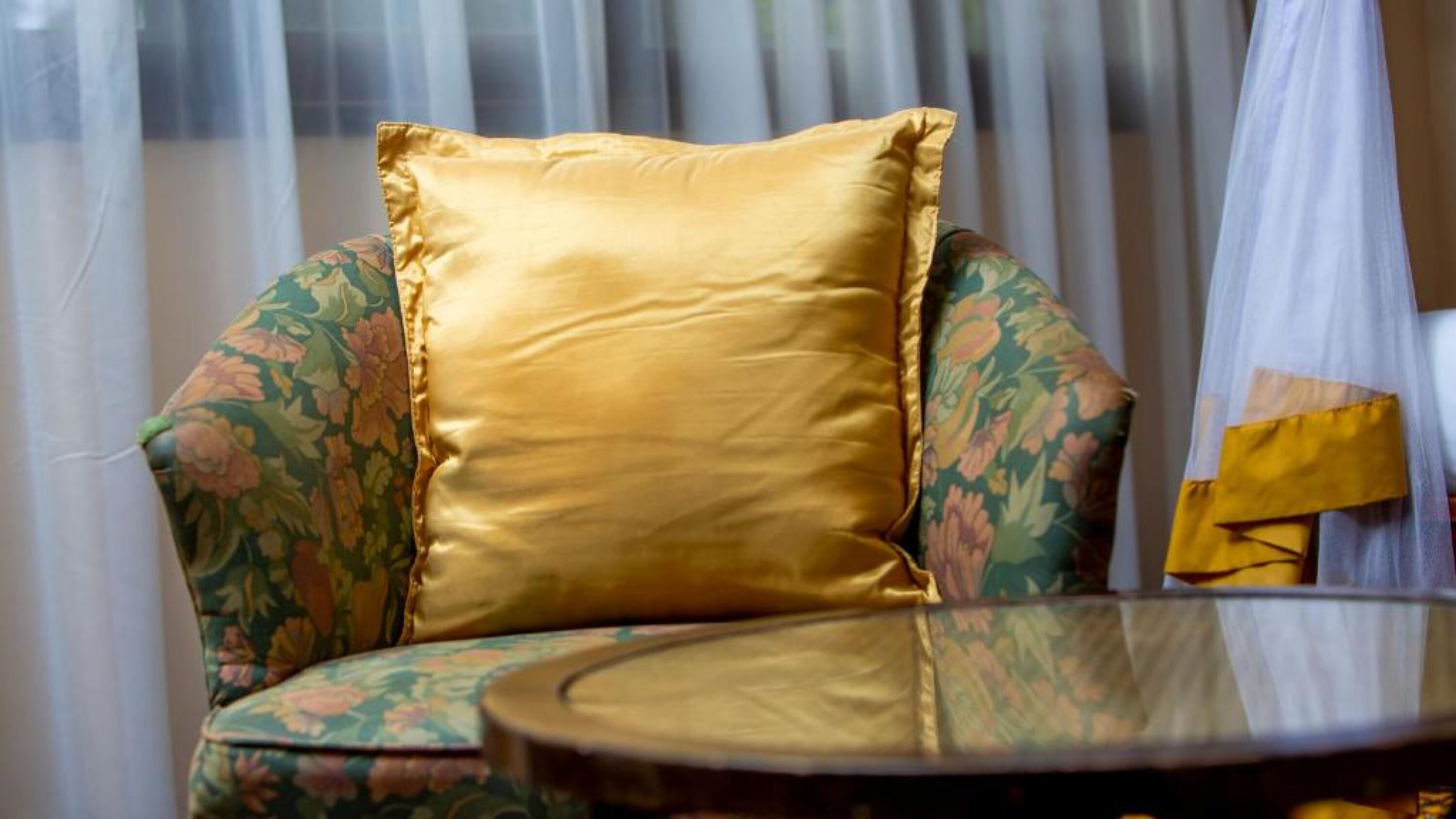 Une chaise à motifs floraux et un coussin doré, placés à côté d’une table en verre.