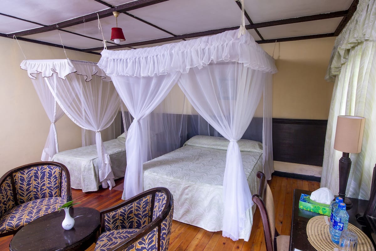 A room with two single beds, each with a canopy bedspread and white curtains. There is a wooden chair with a patterned cushion, a small table with a vase, and a lamp on a side table.