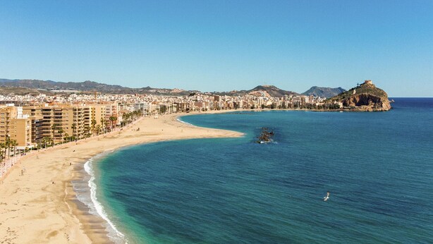 Un paisaje urbano costero con una playa arenosa, mar azul cristalino y una colina destacada con una fortaleza.