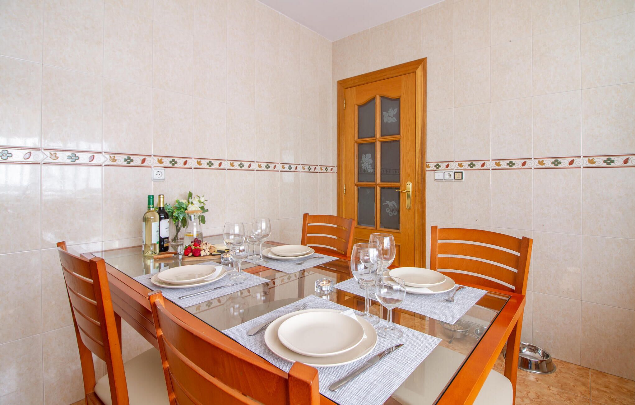 A dining area with a glass table, wooden chairs, and a tiled wall with a decorative border.