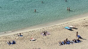 Uma praia com águas cristalinas, pessoas nadando e tomando sol, e edifícios ao fundo.