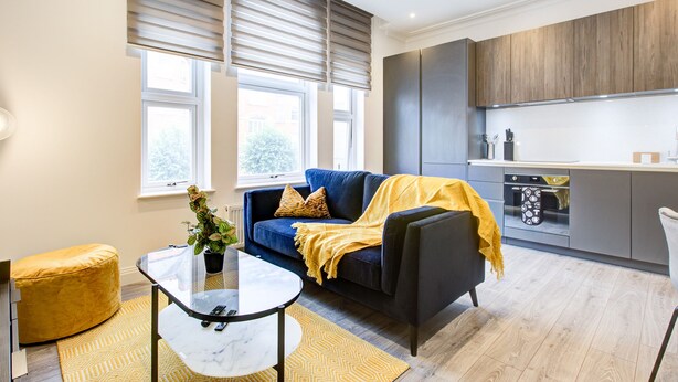 A modern living room with a sofa, a coffee table, and a yellow ottoman. There is a kitchen area with a stove and cabinets.