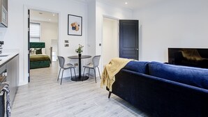 A modern living room with a blue sofa, a dining table with chairs, and a small kitchen area.