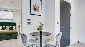 A modern bedroom with a green upholstered bed, a round table with two chairs, and a framed abstract painting on the wall.