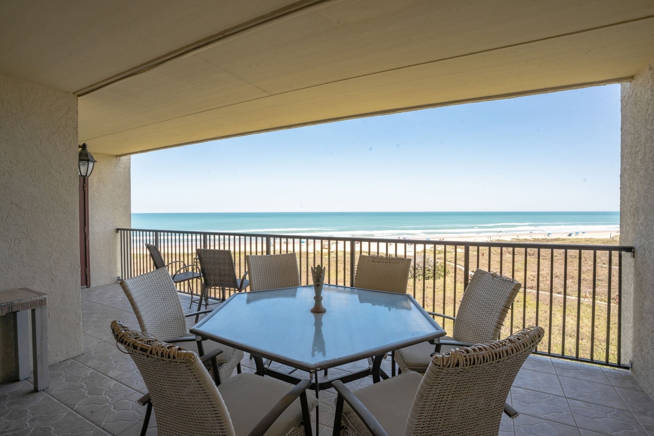 A covered outdoor area with a glass table and wicker chairs, overlooking a beach.