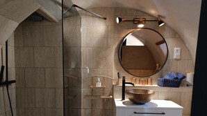 Ein Badezimmer mit einer gläsernen Duschkabine, einem runden Spiegel, einem weißen Waschbecken mit Kupferarmatur und einem weißen Schrank mit Schubladen.