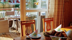 Ein Holztisch mit einer Kaffeetasse, einem Teller mit einem Servietten und einem Brotkorb auf einer Terrasse mit Korbstühlen und Blick auf einen Kanal.
