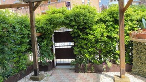 Une pergola en bois, avec un chemin pavé de briques menant à une entrée fermée par un portail, le tout entouré d’une végétation luxuriante et de plantes en pot.