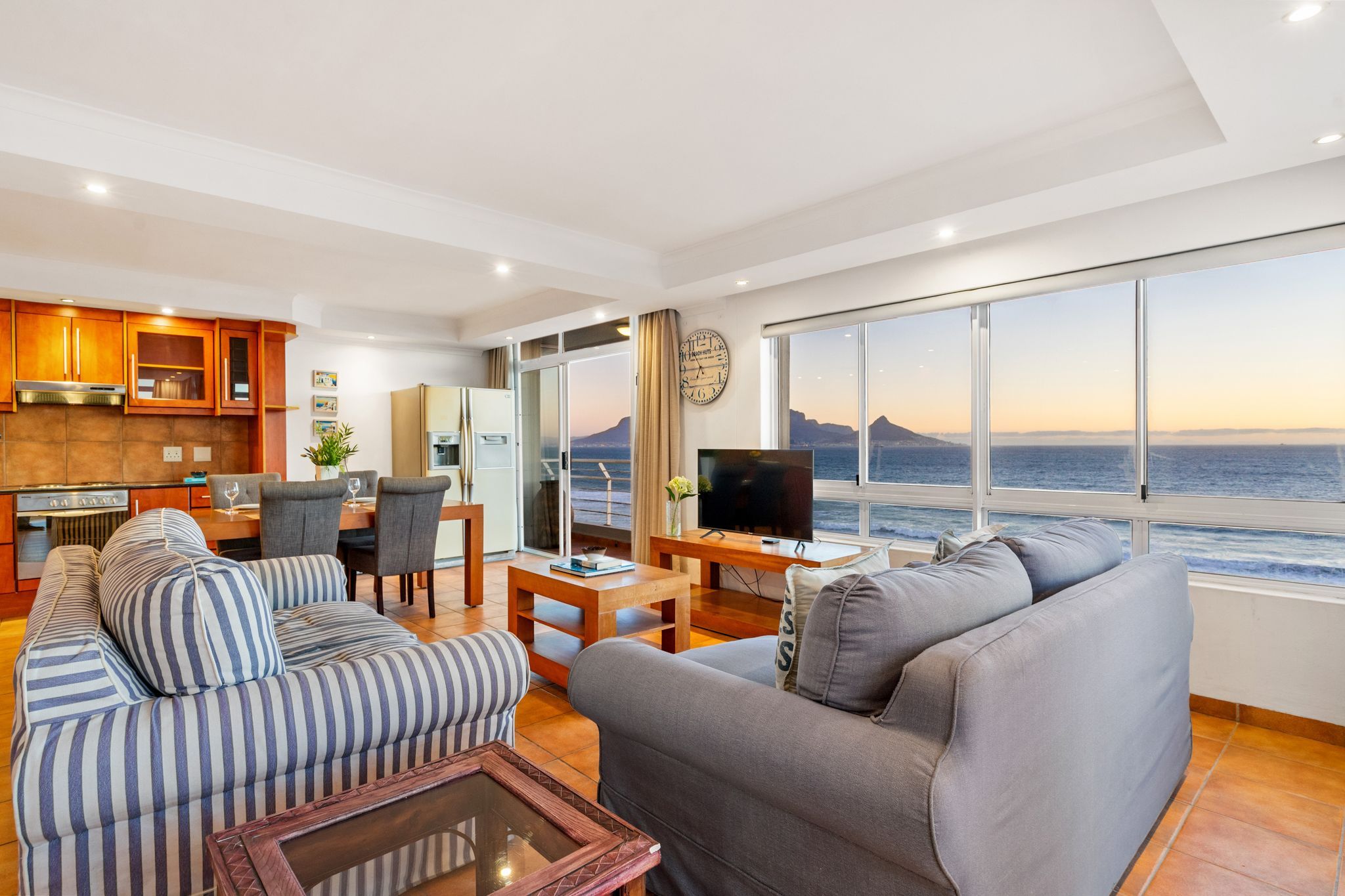 A modern living room with a large TV, comfortable seating, a dining area, and a view of the ocean.
