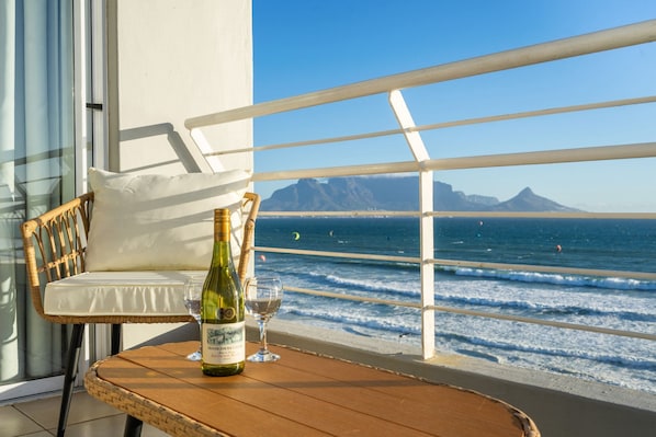A balcony with a wicker chair, a wooden table with a bottle of champagne and a glass, and a view of the ocean and mountains.