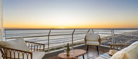 A balcony with wicker furniture, a table with a bottle and glasses, and a view of the ocean and cityscape at sunset.