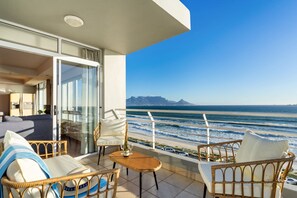A balcony with wicker furniture, a table with a bottle, and a view of the ocean and distant mountains. - Ocean View 701 (Cape Town)