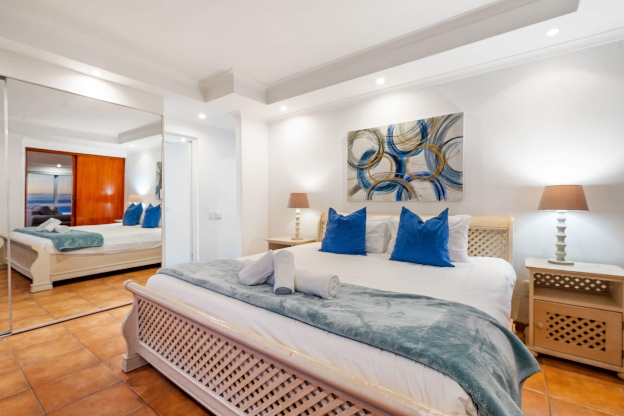 A modern bedroom with a large bed, a mirror, a painting, and a nightstand with a lamp.