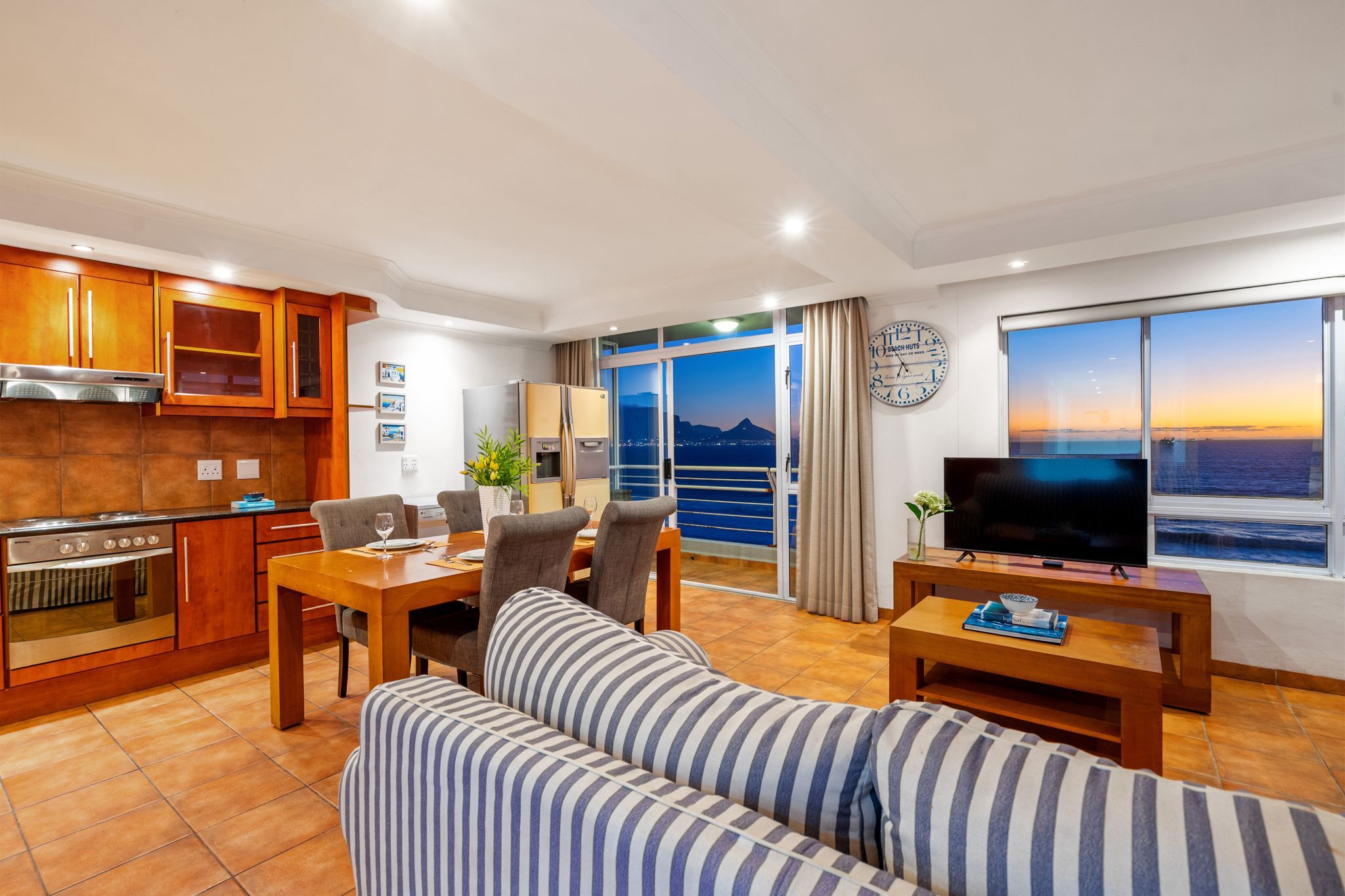 A modern living room with a sofa, dining table, and TV. It features a kitchen with wooden cabinets and a large window with a city view.