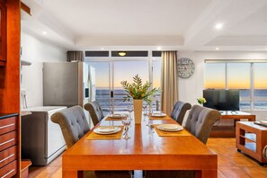 A dining area with a wooden table set for four, a large window with a view of the ocean, and a television on a wooden stand. - Ocean View 701 (Cape Town)