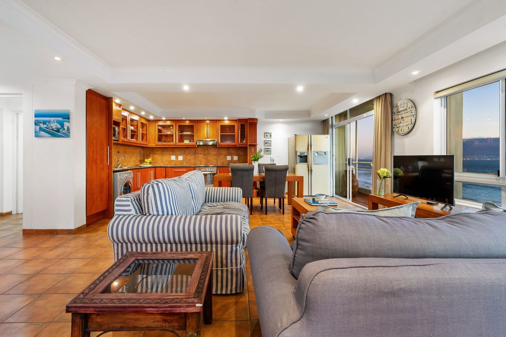 A modern living room with a sofa, a coffee table, and a TV. There is a kitchen with wooden cabinets and a dining area.