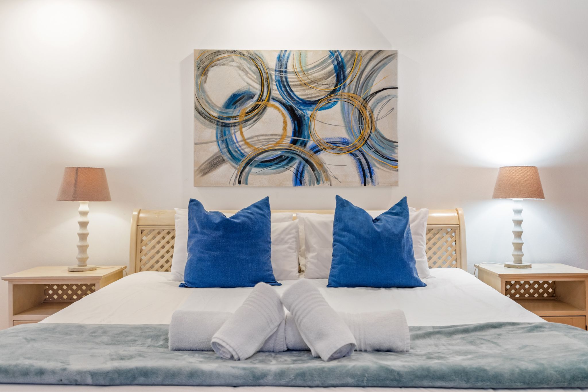 Una cama bien hecha con ropa de cama azul y blanca, dos lámparas de noche y un cuadro abstracto grande en la pared.
