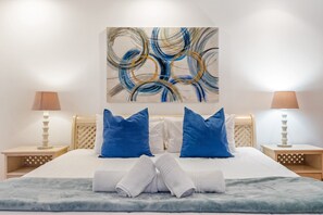A neatly made bed with blue and white bedding, two bedside lamps, and a large abstract painting on the wall. - Ocean View 701 (Cape Town)