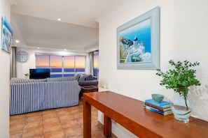 A living room with a sofa, a wooden table, a painting, and a view of the sunset through large windows. - Ocean View 701 (Cape Town)