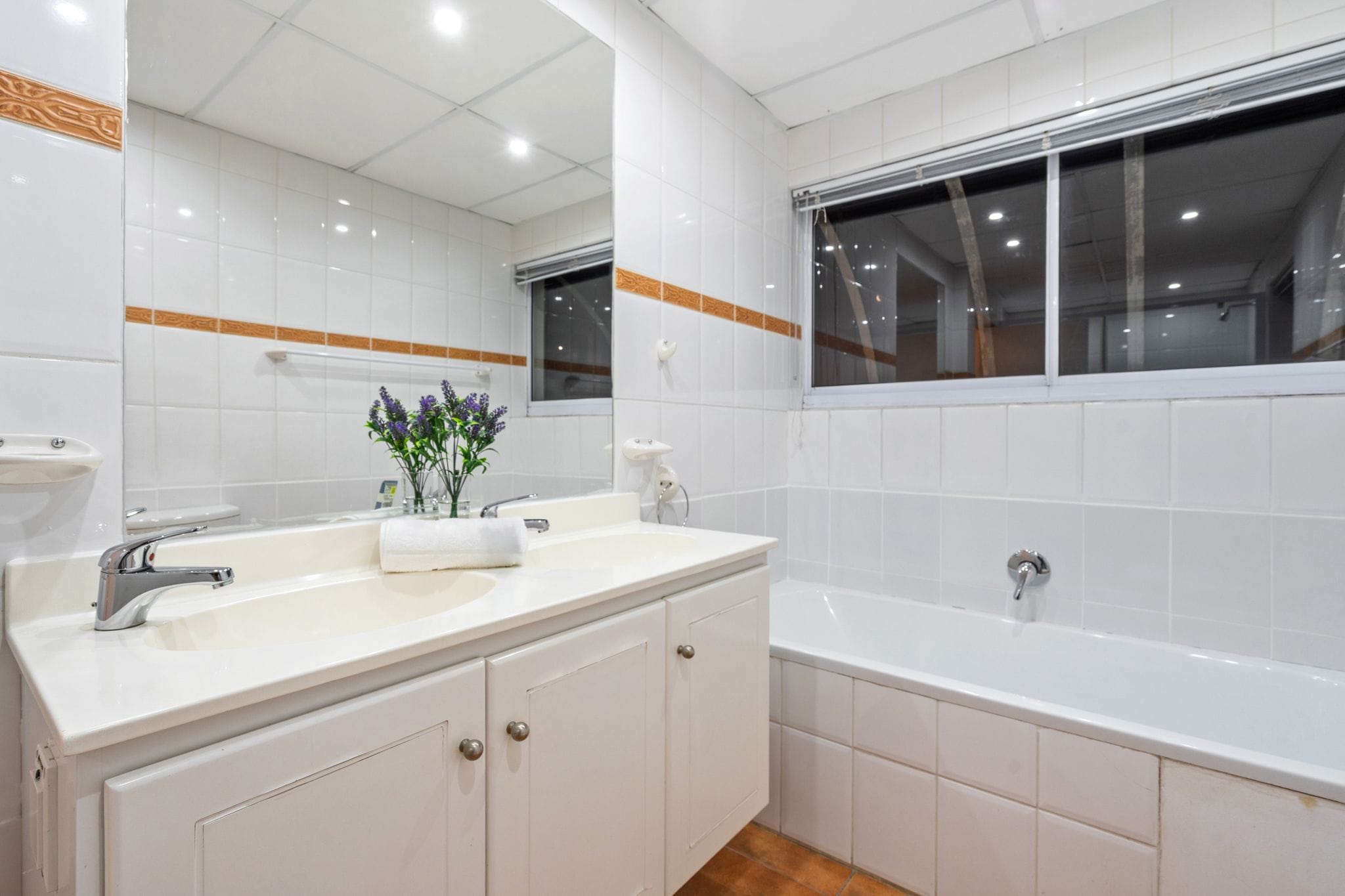 Une salle de bain moderne avec des carreaux blancs, un grand miroir, une fenêtre, un lavabo et une baignoire.