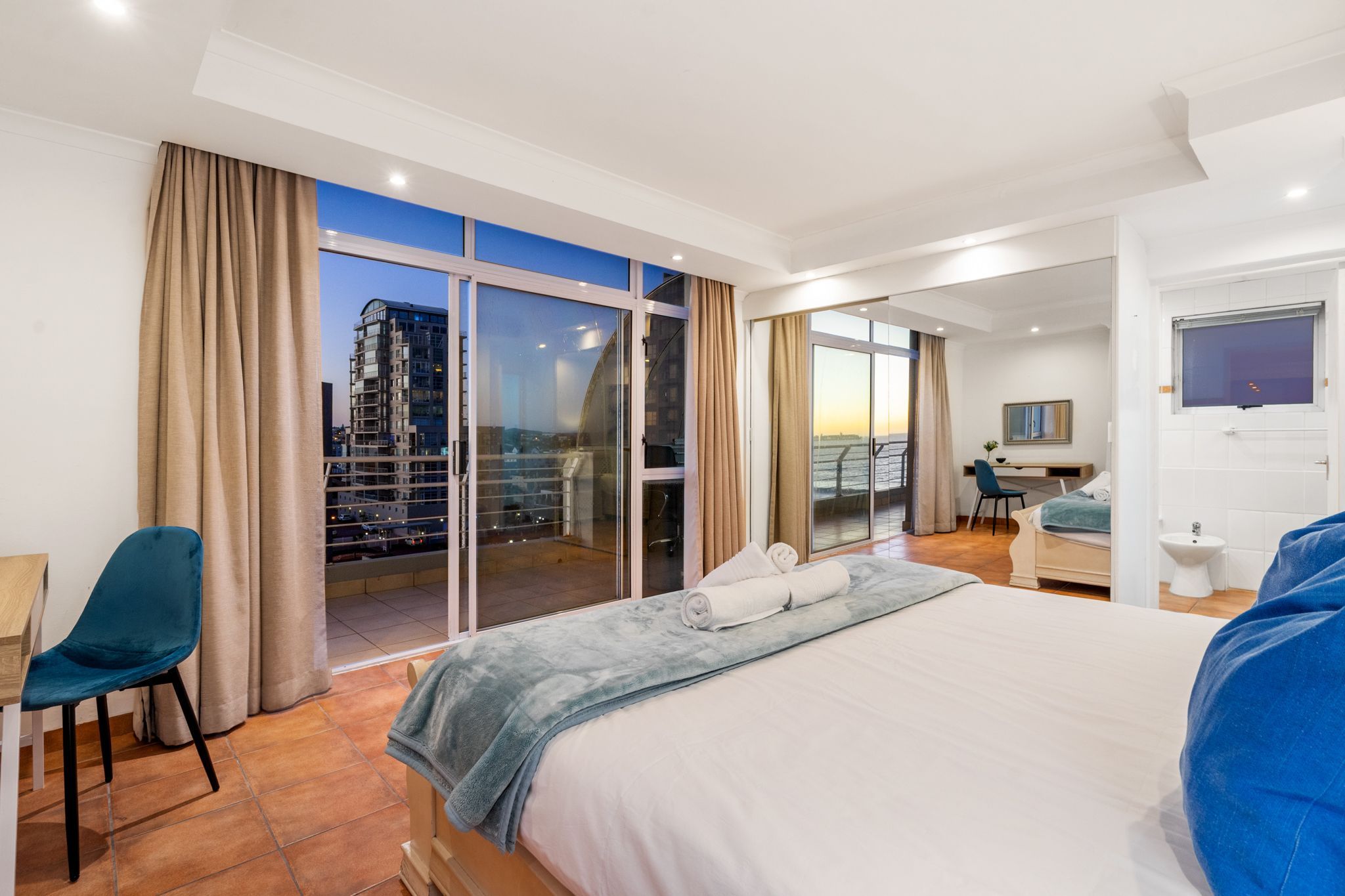 A hotel room with a large bed, a desk, a chair, a balcony with a city view, and a sliding door leading to another room.