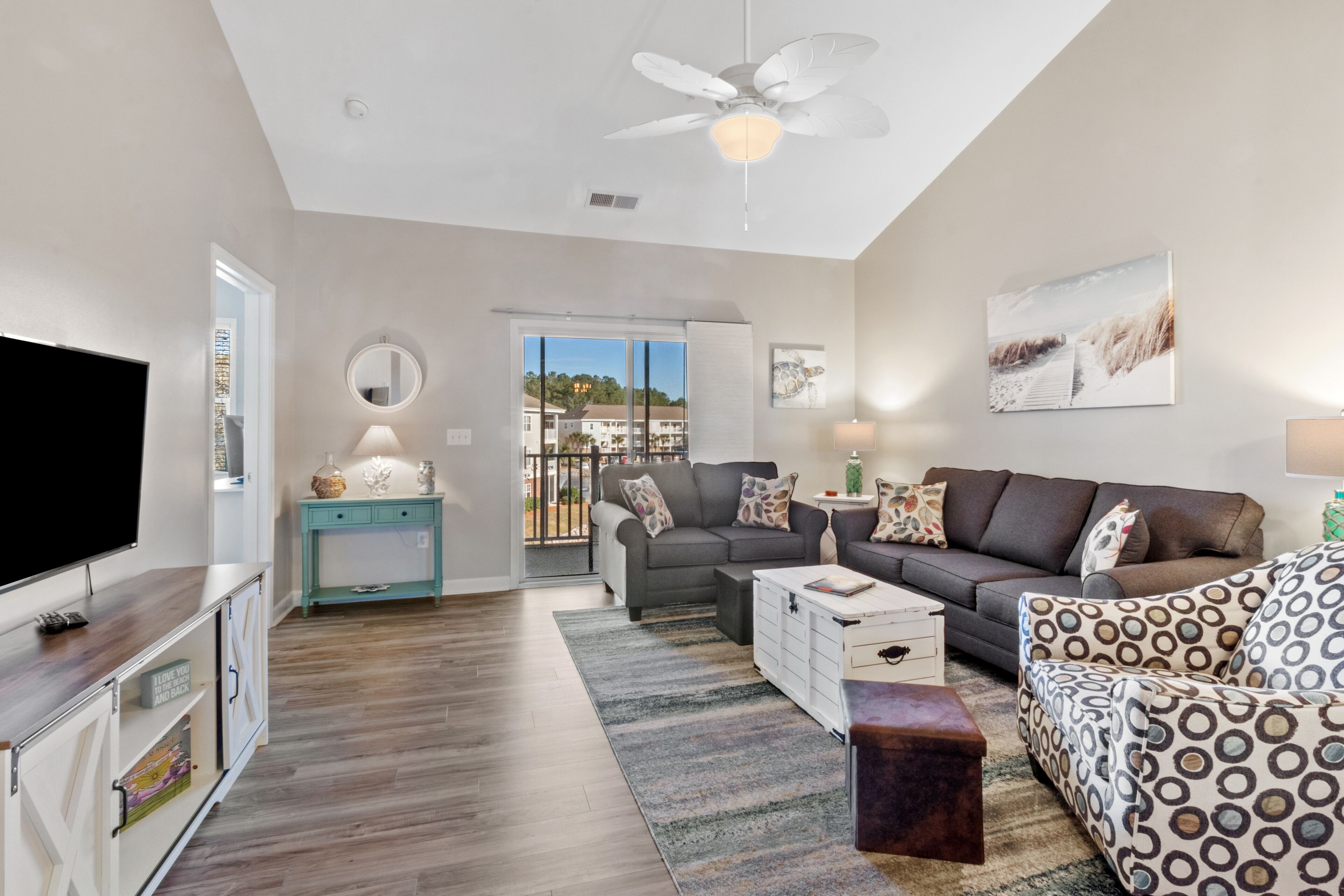 A spacious living room with a large flat-screen TV, a ceiling fan, and a view through a sliding glass door to the outdoors.