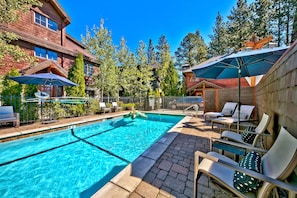 A swimming pool with lounge chairs and umbrellas, surrounded by trees and a building with large windows.