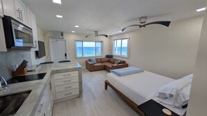 A modern, compact living space with a kitchenette, a bed, a seating area, and large windows offering a view of the sea. - Full Remodel, Direct Beachfront 3rd Floor Corner Unit, Fantastic Views! (Pompano Beach)