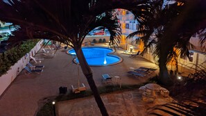 A pool area with lounge chairs and a circular pool, surrounded by palm trees and a building in the background.