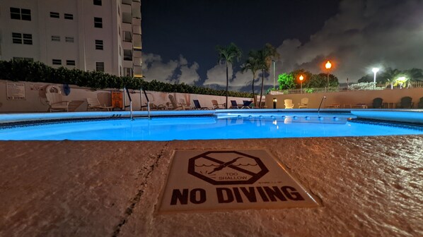 Une zone autour d’une piscine avec un panneau indiquant « INTERDIT DE PLONGER », des chaises longues et un bâtiment en arrière-plan.