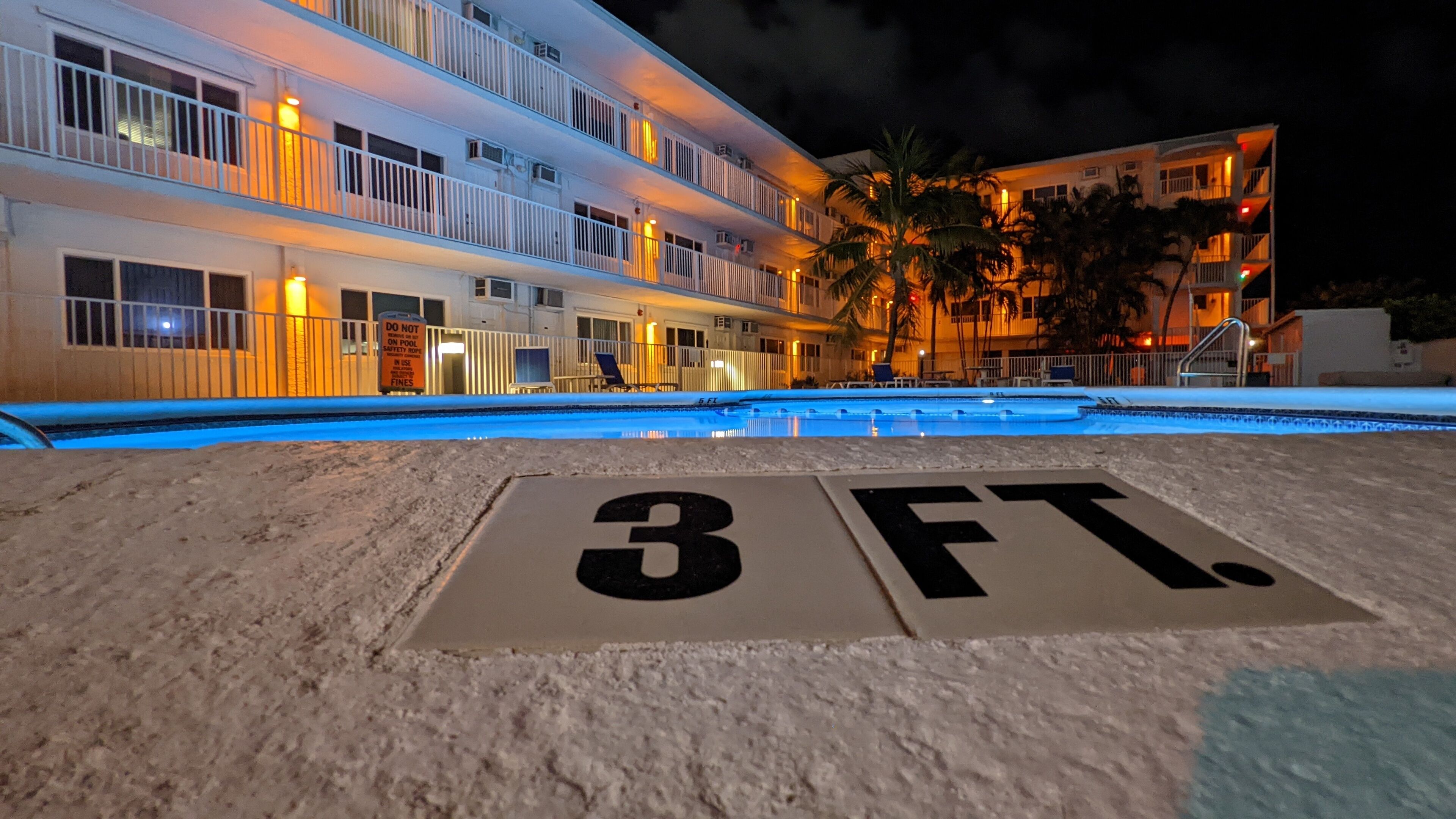 Hotel com piscina, placa indicando a profundidade da piscina e um edifício com várias andares e varandas.