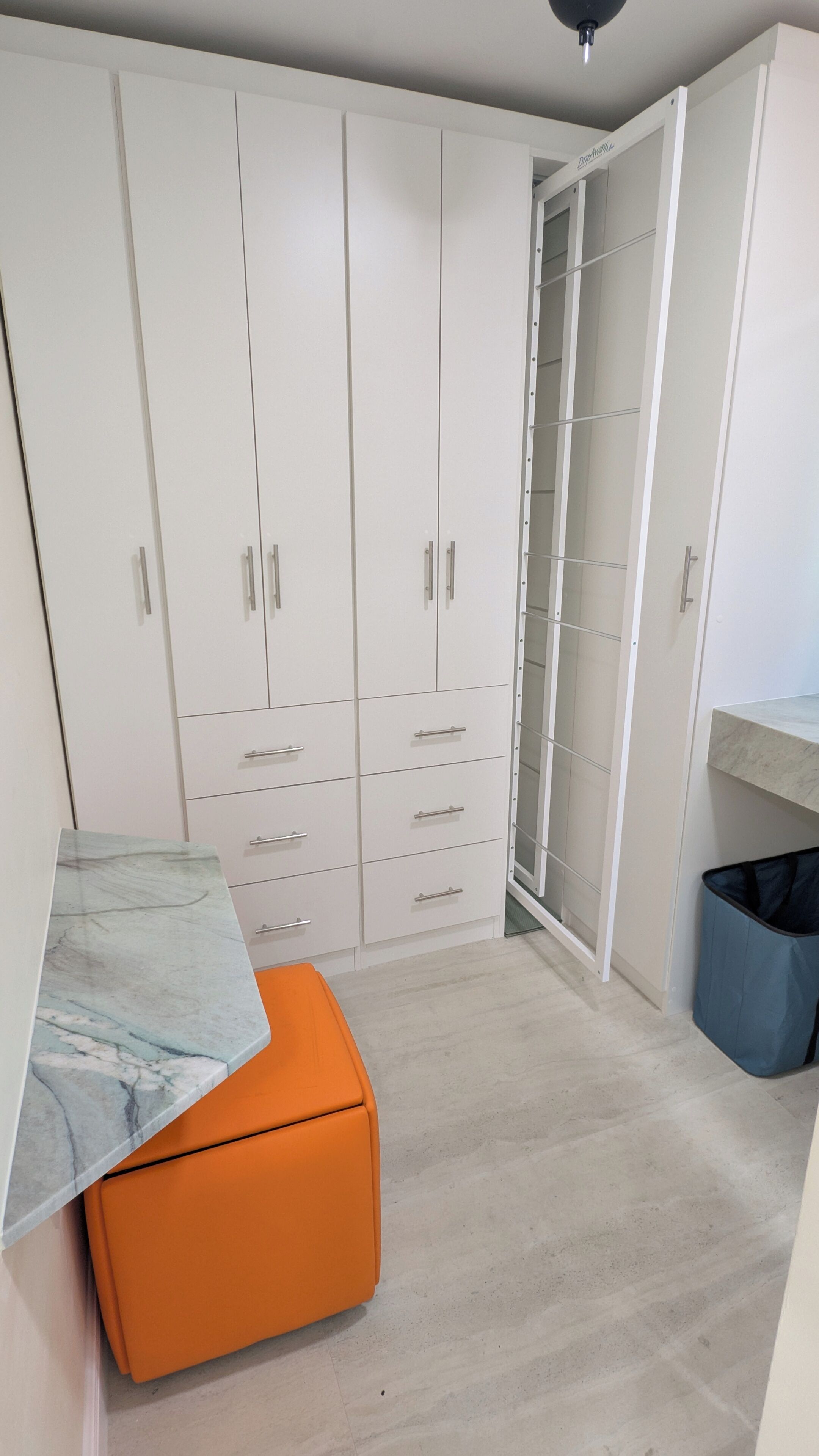A room with a large white wardrobe, a marble-topped table, an orange ottoman, and a blue trash bin.
