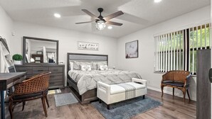 Un dormitorio con cama, dos sillas, escritorio, cómoda, ventilador de techo y ventana con persianas.