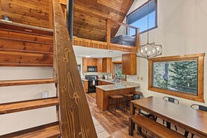 A spacious kitchen with wooden cabinetry and a large island, a dining area with a wooden table and benches, and a lofted sleeping area with a view of trees. - Aspen Acre – Hot tub, wraparound deck, fireplaces & modern mountain charm! (Lead)