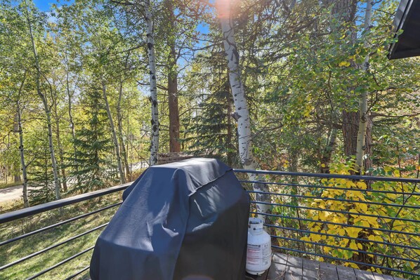 A grill with a protective cover, a propane tank, and a metal railing overlooking a forested area. - Aspen Acre – Hot tub, wraparound deck, fireplaces & modern mountain charm! (Lead)