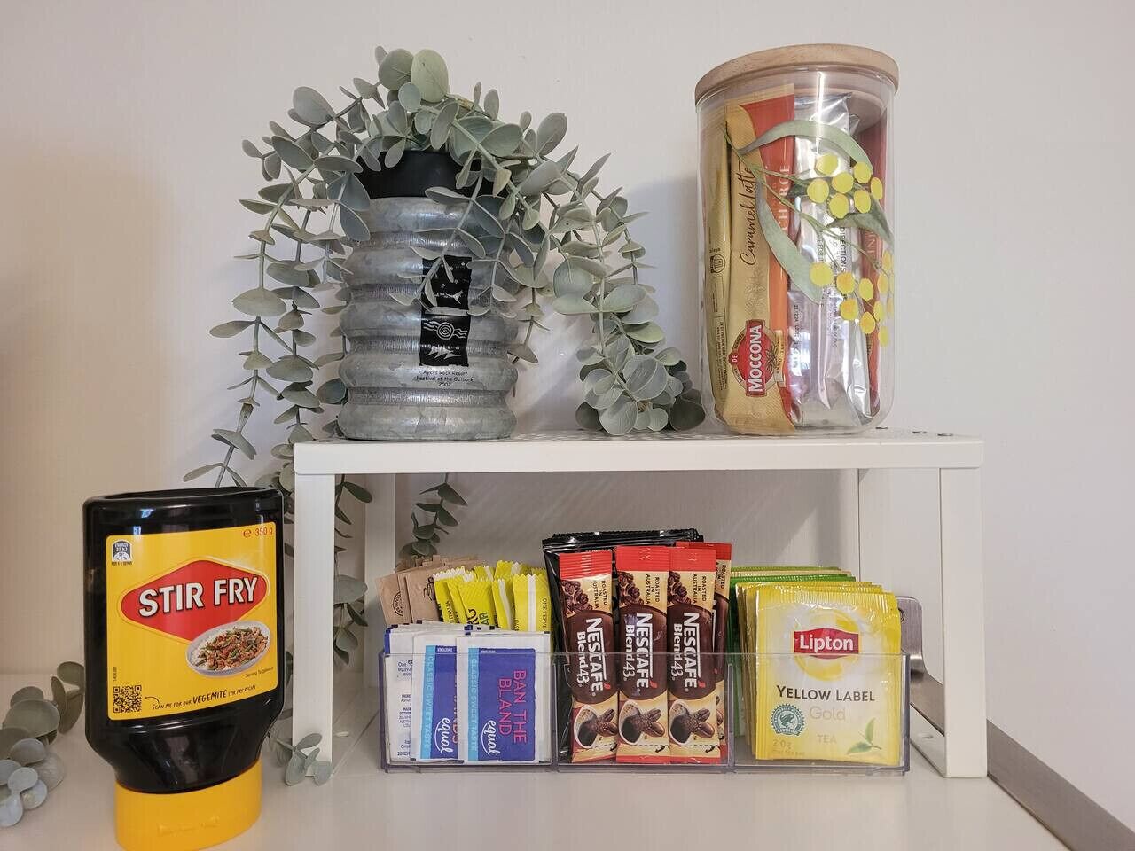 Una estantería con diversos alimentos, incluyendo una botella de salsa para salteado, mezclas de Nescafé, té Lipton y un bote de conservas.