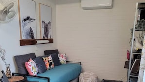 Un salon moderne avec un canapé, une table à manger agrémentée de sets de table en fibres naturelles, un ventilateur sur pied et des œuvres d’art accrochées au mur.
