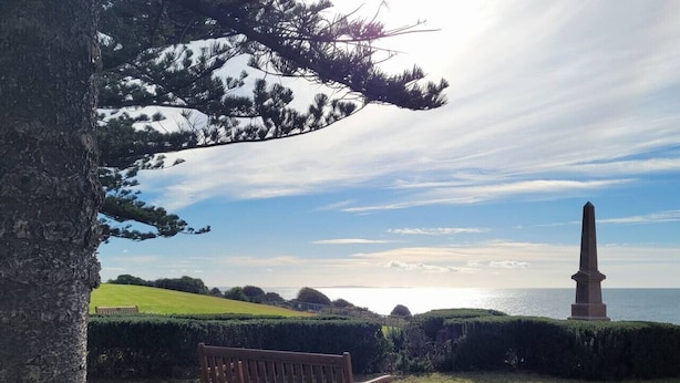 Un banc dans un parc, avec un monument en arrière-plan et une vue sur la mer.