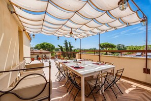 A terrace with a white table set for dining, surrounded by chairs, under a white canopy with hanging lights.