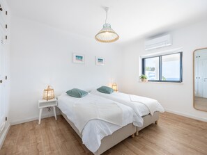 Une chambre à coucher lumineuse et épurée, avec deux lits, une grande fenêtre et un luminaire suspendu.