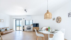 Un vaste espace de vie comprenant un canapé, une table à manger et des chaises. On y trouve également un téléviseur, une lampe en osier et une vue sur l’extérieur grâce à de grandes fenêtres.